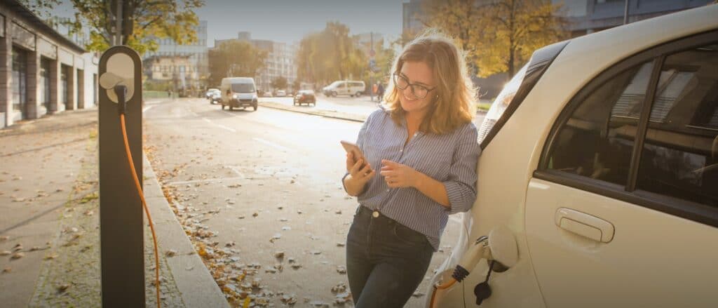 Elektromobilität ganzheitlich gedacht. Titelbild des Beitrags zeigt Frau an Pkw gelehnt vor Ladesäule mit Smartphone in der Hand.